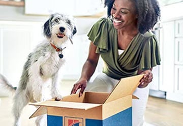 A smiling woman opening a Hill's delivery box on the floor next to her excited scruffy dog.
